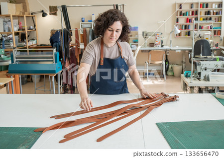Caucasian woman makes belts from genuine leather in a workshop. Caucasian woman makes belts from genuine leather in a workshop. 133656470