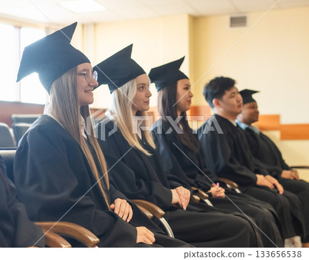 Young people in graduation gowns listen to a speech. Young people in graduation gowns listen to a speech. 133656538