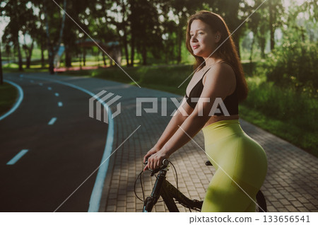 A Caucasian woman in yellow leggings and a crop top rides a bicycle in tights in the park.  133656541