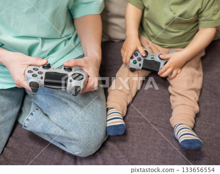 Caucasian woman teaches her little son to play console. Joystick in hands. Caucasian woman teaches her little son to play console. Joystick in hands. 133656554