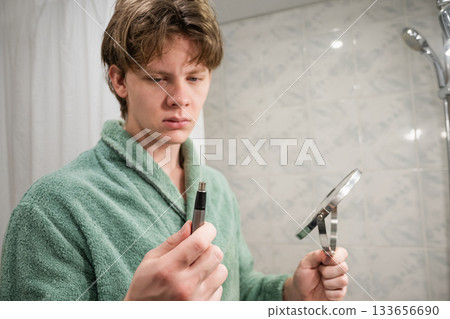 Young caucasian man wearing green bathrobe uses an electric trimmer to groom his facial hair. Morning Shaving Routine. Close up. 133656690