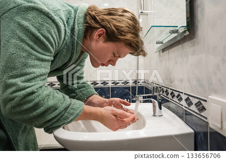 Young man in bathrobe washing his face with water over bathroom sink as part of his morning hygiene and skincare routine. Side view. Young man in bathrobe washing his face with water over bathroom sink as part of his morning hygiene and skincare routine. Side view. 133656706