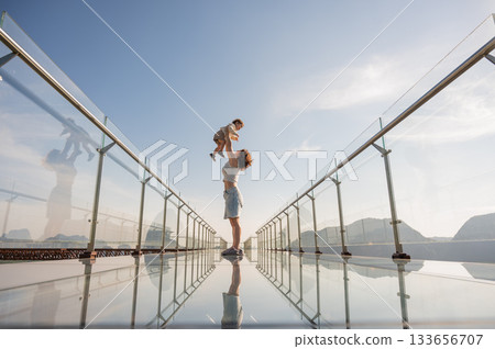 Caucasian woman holds her little son in her arms while standing on a glass bridge in the mountains. Caucasian woman holds her little son in her arms while standing on a glass bridge in the mountains. 133656707
