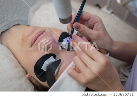 A woman undergoing eyelash extension procedure using an ultraviolet lamp. A woman undergoing eyelash extension procedure using an ultraviolet lamp. 133656751