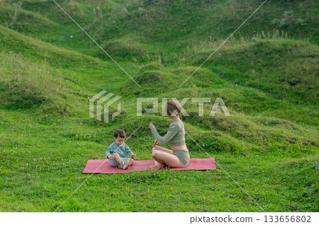 Caucasian woman doing yoga with her little son outdoors. Caucasian woman doing yoga with her little son outdoors. 133656802