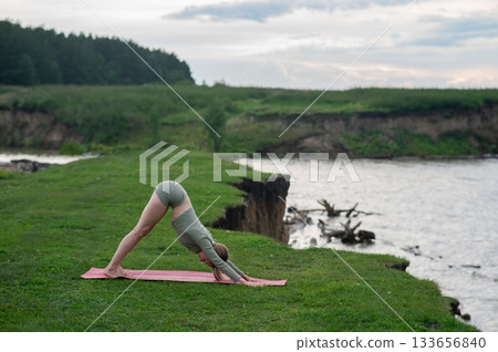 Caucasian woman doing yoga on the river bank. Downward facing dog pose. Caucasian woman doing yoga on the river bank. Downward facing dog pose. 133656840