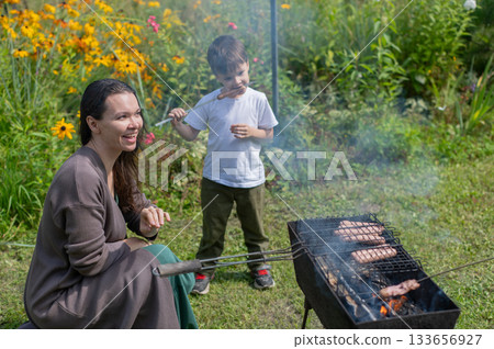 Caucasian woman grilling sausages on a barbecue with her son.  133656927