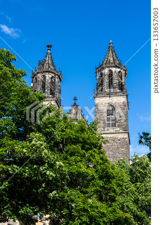Majestic Magdeburg Dom towers rise above lush green trees under vibrant blue sky. Gothic stone details, pointed spires and central clock create striking contrast with bright summer atmosphere Majestic Magdeburg Dom towers rise above lush green trees under vibrant blue sky. Gothic stone details, pointed spires and central clock create striking contrast with bright summer atmosphere 133657003