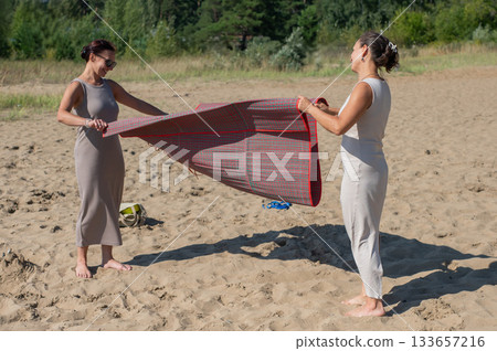 Two Caucasian women spread a blanket on the beach for a picnic. Two Caucasian women spread a blanket on the beach for a picnic. 133657216