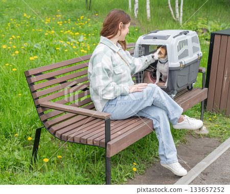 Caucasian woman traveling with dog. Owner sits on bench with Jack Russell Terrier in cage for safe travel. 133657252