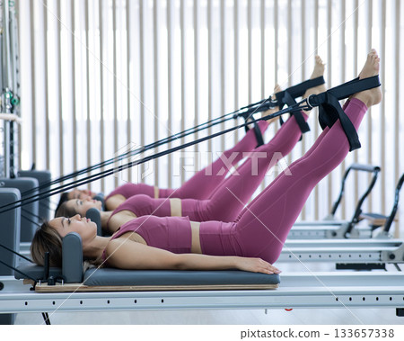 Three Asian women in pink sportswear doing pilates exercises with a reformer bed. Three Asian women in pink sportswear doing pilates exercises with a reformer bed. 133657338