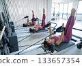 Three Asian women in pink sportswear doing pilates exercises with a reformer bed.  133657354