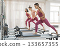 Three Asian women in pink sportswear doing pilates exercises with a reformer bed.  133657355