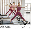 Three Asian women in pink sportswear doing pilates exercises with a reformer bed.  133657359