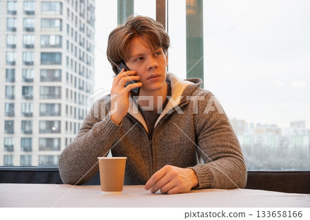Young caucasian man sitting in cafe near window, talking on smartphone and drinking coffee during casual break in an urban setting. Coffee break. Young caucasian man sitting in cafe near window, talking on smartphone and drinking coffee during casual break in an urban setting. Coffee break. 133658166