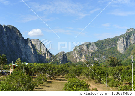 Vast limestone mountains dominate the landscape, with lush green trees and a small cluster of buildings in the foreground. A clear blue sky with sparse clouds completes the scenic view. Vast limestone mountains dominate the landscape, with lush green trees and a small cluster of buildings in the foreground. A clear blue sky with sparse clouds completes the scenic view. 133658499