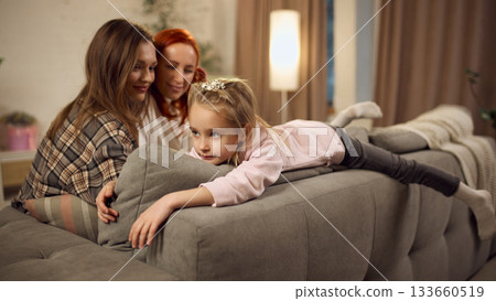 Little girl lying on sofa while her two mothers watch her with affection 133660519