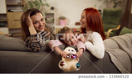 Smiling child leaning over sofa between her two moms in cozy family setting 133660520