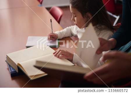 Young Student Writing in Class While Teacher Assists, Surrounded by Books and Notebooks 133660572