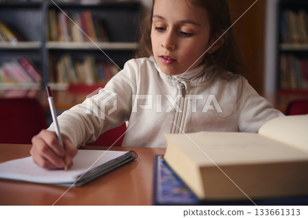 Focused Girl Writing in Notebook at Library Desk with Open Books Nearby Focused Girl Writing in Notebook at Library Desk with Open Books Nearby 133661313