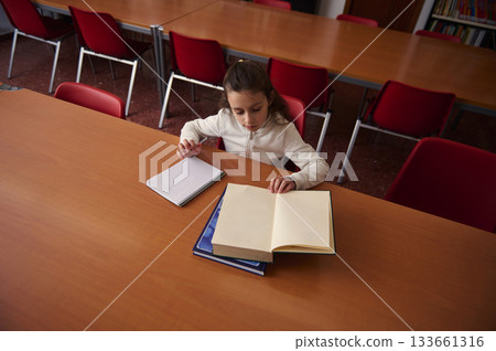 Girl Studying at Library Desk With Open Book and Notebooks in Quiet School Setting Girl Studying at Library Desk With Open Book and Notebooks in Quiet School Setting 133661316
