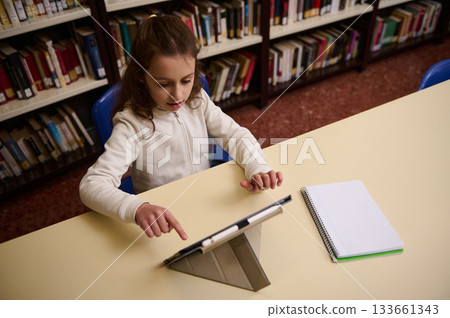 Young Girl Studying in Library Using Tablet With Notebook on Desk 133661343