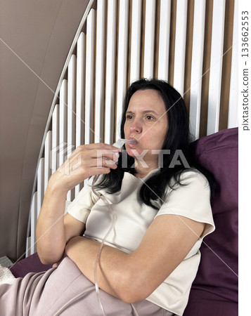Adult brunette woman inhaling medication through a portable nebulizer device in a bedroom environment, home respiratory care, chronic illness management, realistic medical lifestyle photo 133662553