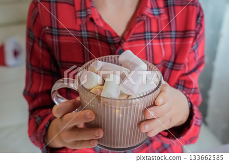 Close-up photo of a person holding a mug of hot cocoa with marshmallows near the chest. No face visible. Warm winter atmosphere, cozy mood, holiday theme, soft neutral background. 133662585