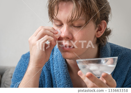 Young caucasian man sitting on sofa in bathrobe applying ice cubes to face during refreshing home skincare routine. Selfcare Routine. Close up. Young caucasian man sitting on sofa in bathrobe applying ice cubes to face during refreshing home skincare routine. Selfcare Routine. Close up. 133663358