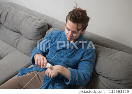Young caucasian man in blue bathrobe applying skincare cream with under-eye patches while sitting on sofa during home selfcare routine. Mens Home Self-Care Routine Young caucasian man in blue bathrobe applying skincare cream with under-eye patches while sitting on sofa during home selfcare routine. Mens Home Self-Care Routine 133663359