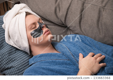 Young caucasian man lying on sofa with white towel on his head and black facial mask applied to his skin during relaxation skincare session. Mens Home Self-Care Routine Young caucasian man lying on sofa with white towel on his head and black facial mask applied to his skin during relaxation skincare session. Mens Home Self-Care Routine 133663368