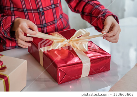 Close-up of hands tying a bow on a red gift box. Face not visible. Festive holiday scene. 133663602