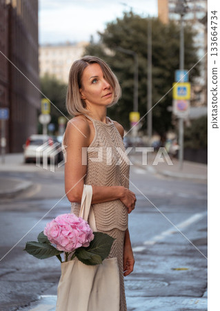 A woman stands on a city street in soft evening light, holding a canvas tote with a pink hydrangea, muted beige tones creating a calm mood A woman stands on a city street in soft evening light, holding a canvas tote with a pink hydrangea, muted beige tones creating a calm mood 133664734