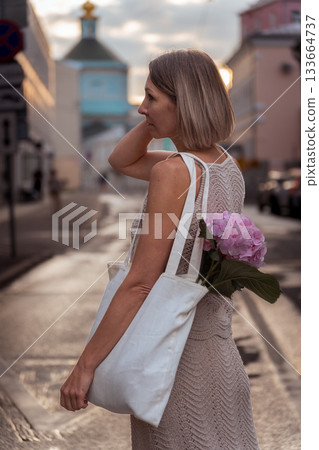 A woman walks along a sunlit city street with a canvas tote and pink hydrangea, soft warm tones creating a calm evening atmosphere A woman walks along a sunlit city street with a canvas tote and pink hydrangea, soft warm tones creating a calm evening atmosphere 133664737