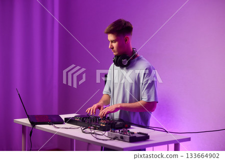 Young DJ with headphones, Skilled DJ engaging with equipment in lively environment, Energetic DJ prepping for live performance amid colorful lighting and fashionable attire Young DJ with headphones, Skilled DJ engaging with equipment in lively environment, Energetic DJ prepping for live performance amid colorful lighting and fashionable attire 133664902