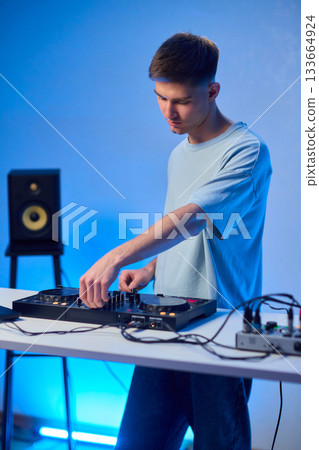 Young DJ with headphones, Skilled DJ engaging with equipment in lively environment, Energetic DJ prepping for live performance amid colorful lighting and fashionable attire Young DJ with headphones, Skilled DJ engaging with equipment in lively environment, Energetic DJ prepping for live performance amid colorful lighting and fashionable attire 133664924