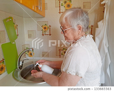 An elderly woman washes her hands with soap under running water from a faucet in a kitchen sink. An elderly woman washes her hands with soap under running water from a faucet in a kitchen sink. 133665563