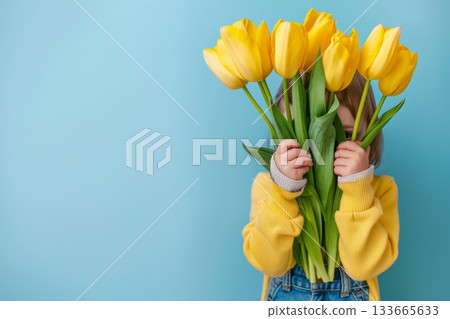 A joyful child peeks from behind a cheerful bunch of yellow tulips, showcasing a playful spirit. The soft blue background enhances the vibrant colors A joyful child peeks from behind a cheerful bunch of yellow tulips, showcasing a playful spirit. The soft blue background enhances the vibrant colors 133665633