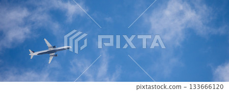 Commercial airplane flying above light clouds with clear view of the aircraft underside against a bright blue sky 133666120