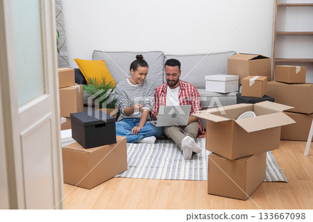 A couple, amidst boxes in their new home, utilizes a laptop for interior design choices, online shopping, or moving arrangements 133667098