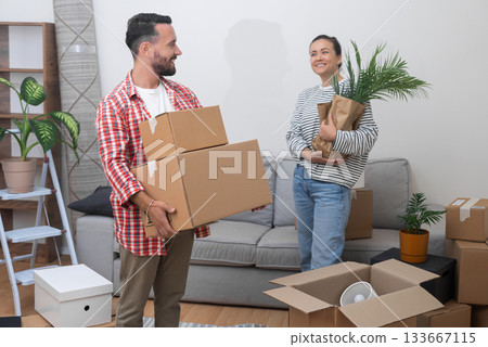 Settling In Abroad: A content and delighted couple, after relocating internationally, stands in their new living room with a pile of cardboard relocation service boxes, feeling at home. 133667115