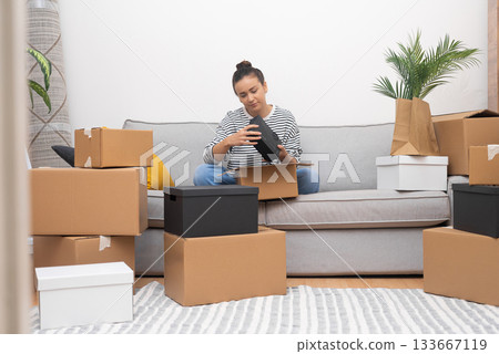Relocation takes center stage as a young woman sits on the sofa in her new home, unpacking boxes filled with belongings on moving day, emphasizing the real estate experience. Relocation takes center stage as a young woman sits on the sofa in her new home, unpacking boxes filled with belongings on moving day, emphasizing the real estate experience. 133667119