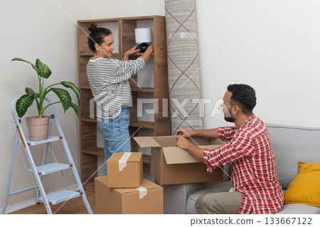 cheerful family, husband and wife, unpacks and arranges a lamp on an empty shelf in their new home, savoring the special moments of their relocation cheerful family, husband and wife, unpacks and arranges a lamp on an empty shelf in their new home, savoring the special moments of their relocation 133667122