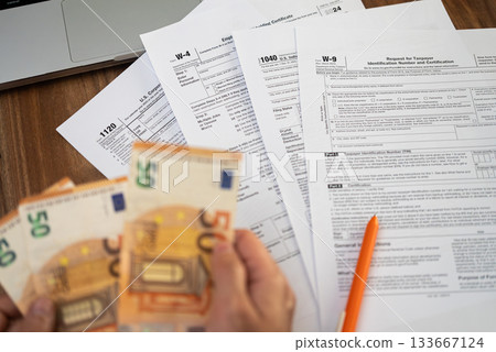 Close-up of hands holding euro bills over various tax forms including W-4, 1040, W-9, and 1120. Tax season, financial accounting, and tax refund concept Close-up of hands holding euro bills over various tax forms including W-4, 1040, W-9, and 1120. Tax season, financial accounting, and tax refund concept 133667124