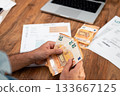 Close-up of hands holding euro banknotes over utility bills and laptop on wooden desk. Concept of personal finance, managing household expenses, and budgeting  133667125