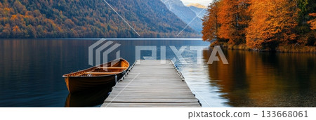 Wooden rowing boat moored to a wooden pier on a tranquil lake surrounded by colorful autumn foliage and majestic mountains, creating a picturesque and serene landscape 133668061