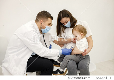 Male doctor in white medical gown examining boy with stethoskope. Boys and woman wearing white t-shirts. Doctor and mother wearing face masks. 133668194