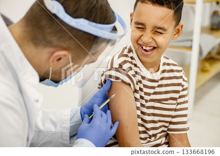 Male doctor is doing a vaccination. Man wearing white medical gown. Boy wearing brown plaid t-shirt. Male doctor is doing a vaccination. Man wearing white medical gown. Boy wearing brown plaid t-shirt. 133668196