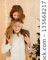 Portrait of a little girl celebrating Christmas with her mom at home. Girl and her mother having fun while standing near Christmas tree. Family wearing beige knitted clothes. 133668217