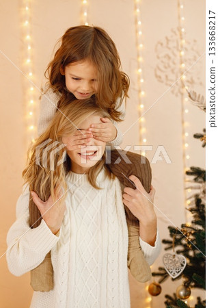 Portrait of a little girl celebrating Christmas with her mom at home. Girl and her mother having fun while standing near Christmas tree. Family wearing beige knitted clothes. Portrait of a little girl celebrating Christmas with her mom at home. Girl and her mother having fun while standing near Christmas tree. Family wearing beige knitted clothes. 133668217